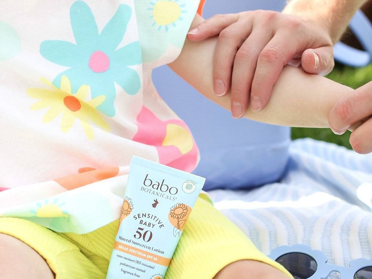 Hands apply sunscreen to a child's arm beside the blue tube outdoors.