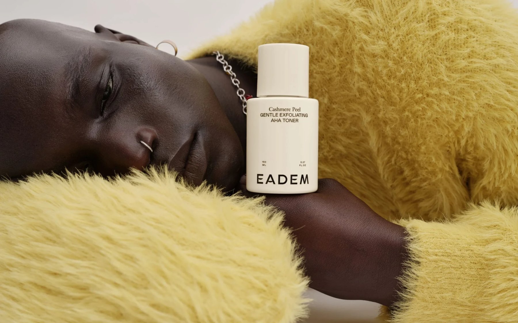 Model in yellow knit holds Eadem bottle near face.