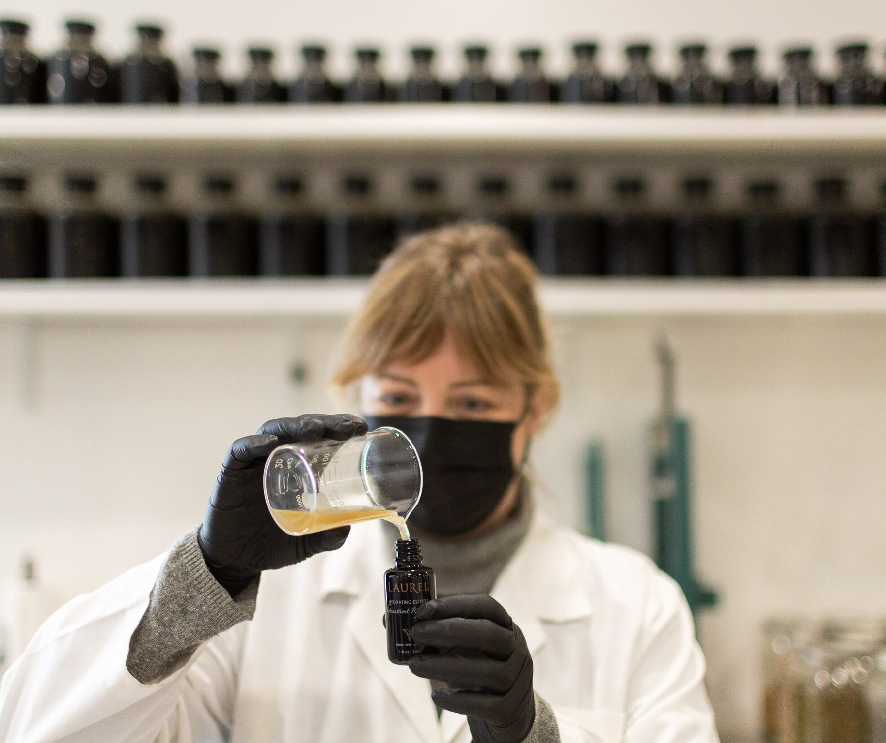 A masked lab worker fills a Laurel bottle from a beaker.