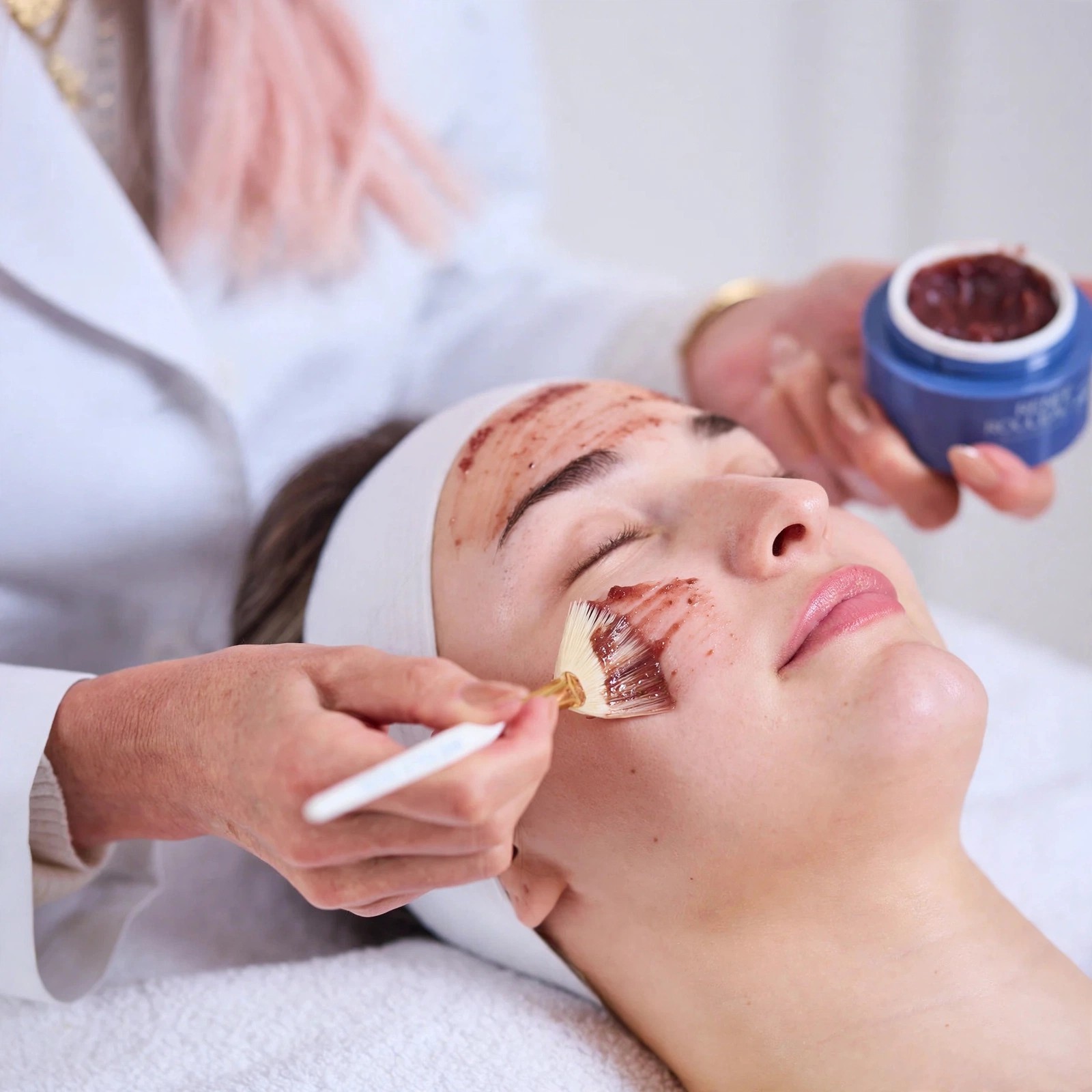 An esthetician brushes berry colored peel onto a reclining client.