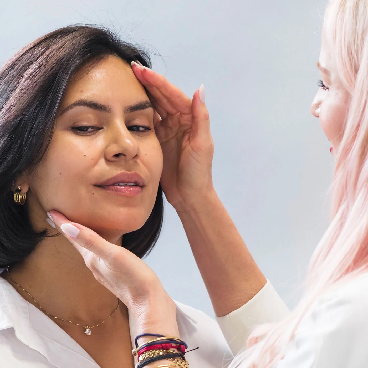 A pink haired esthetician holds a clients face for skin consultation.