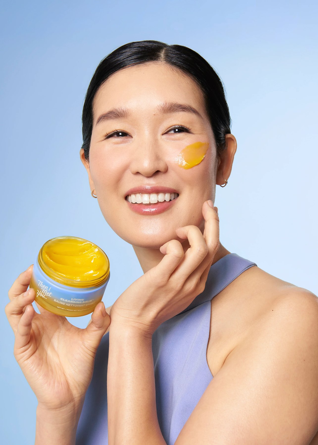 Woman holding open yellow balm with swatch on her cheek.