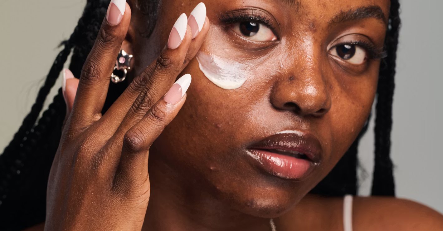 A woman applies cream to her cheek with a fingertip.