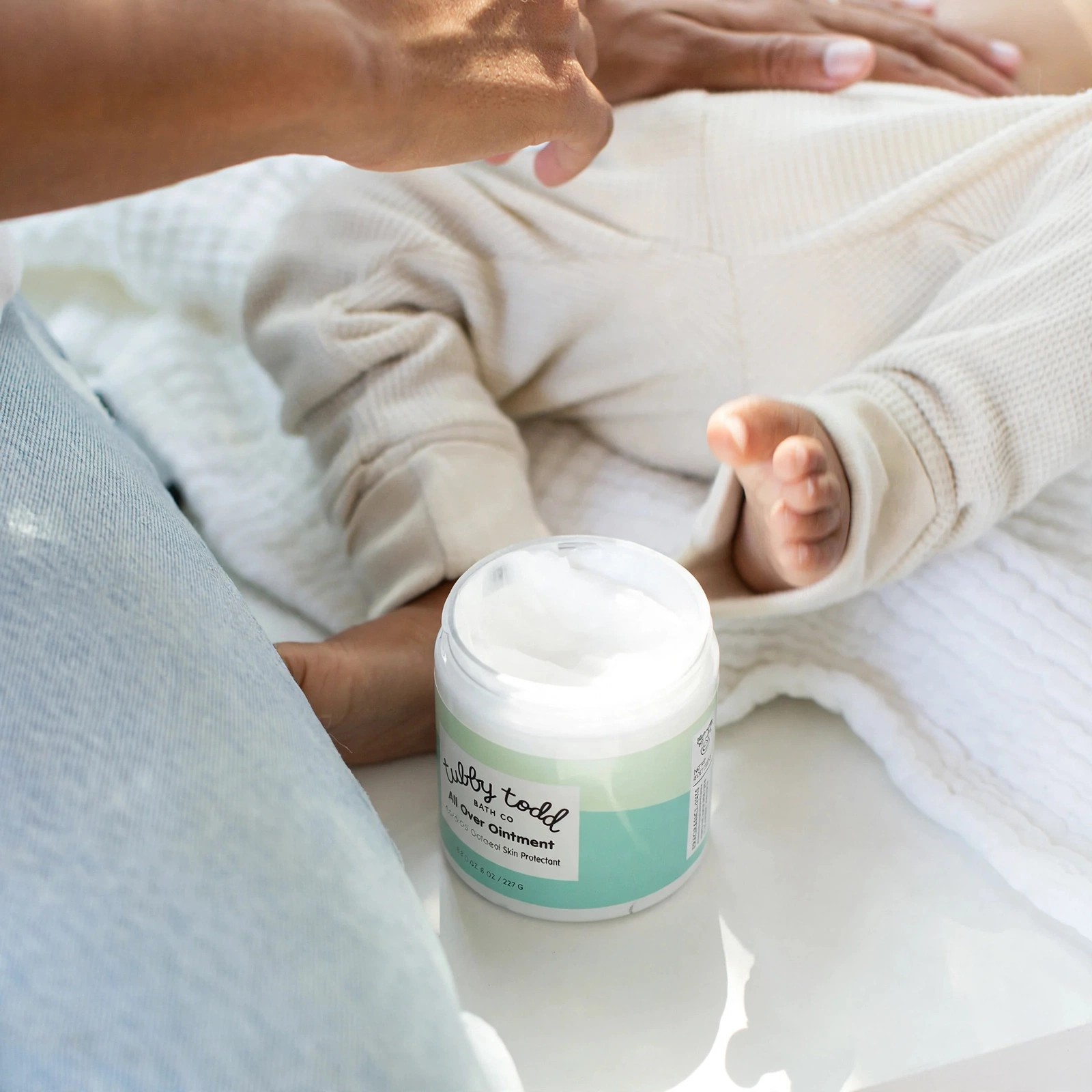 Open jar of All Over Ointment beside a baby on bedding.