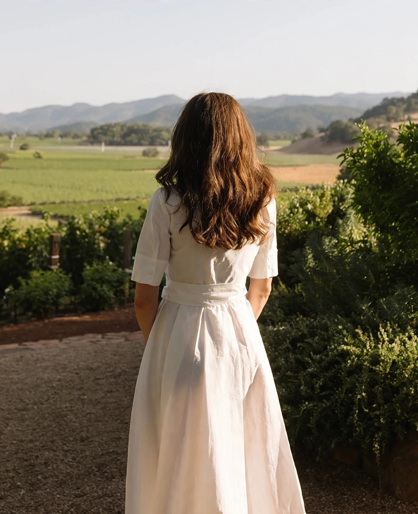 Woman in white dress gazes across vineyard hills.